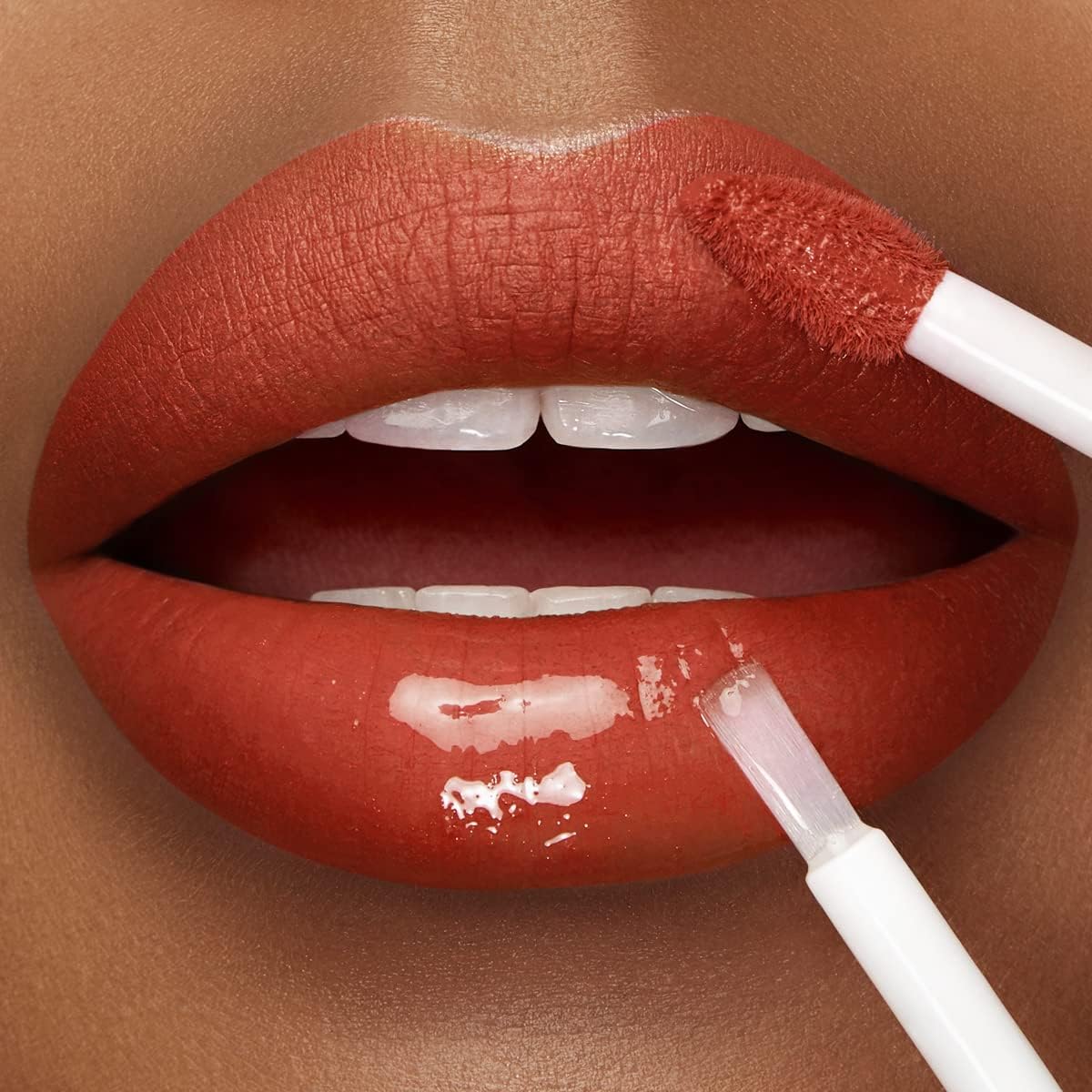 Close-up of a person's lips wearing a matte terracotta red lipstick, with a glossy applicator applying a clear lip gloss to the lower lip.