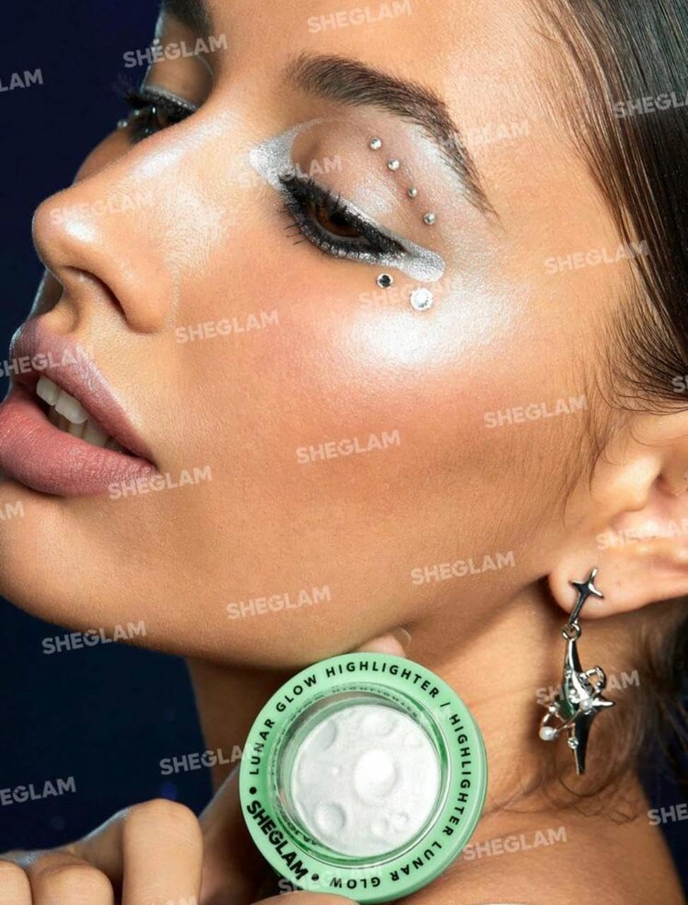 Close-up of a woman's face with dramatic eye makeup and rhinestone embellishments, holding a green-lidded jar of SHEGLAM Glitter Glow Highlighter near her cheek. The image has a dark blue background and multiple SHEGLAM watermarks.