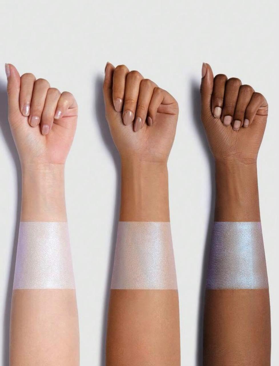 Three human forearms shown side by side against a light gray background, each with a horizontal strip of iridescent shimmering product applied across the forearm. The skin tones range from very light beige to deep brown, and the shimmering bands appear in pale pink, sheer nude, and silvery lavender tones.